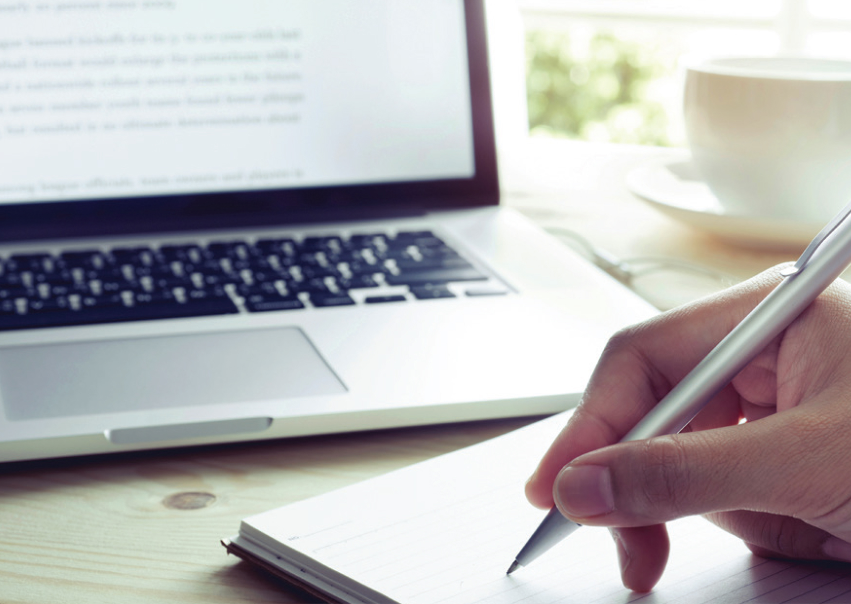 Person writing on notepad; computer in background